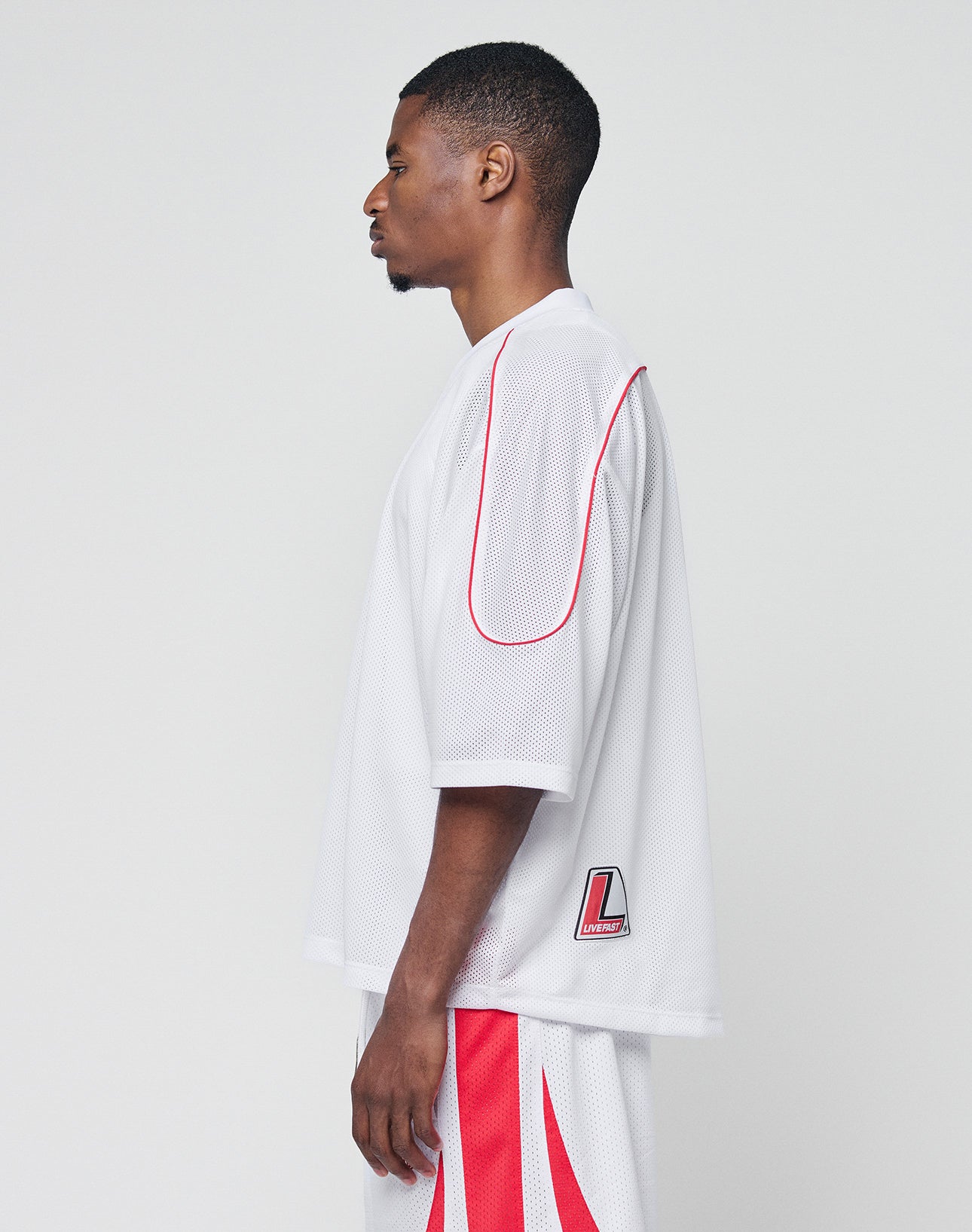 A man stands in profile against a plain background, wearing the LIVE FAST DIE YOUNG Basic Mesh Jersey with red trim and V-neck, paired with matching white and red athletic shorts for an oversized look.
