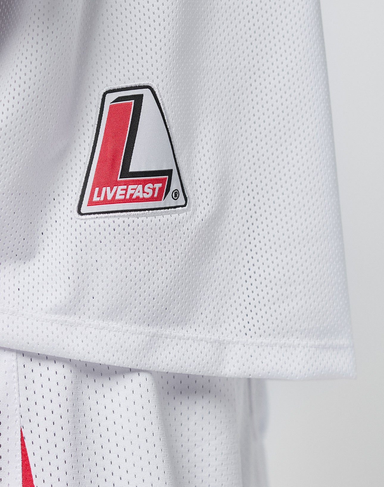 A close-up of the Basic Mesh Jersey by LIVE FAST DIE YOUNG, featuring a white mesh fabric, V-Ausschnitt, red and black L logo, and LIVE FAST text on the upper left. Made from 100% polyester with an oversize fit for comfort.