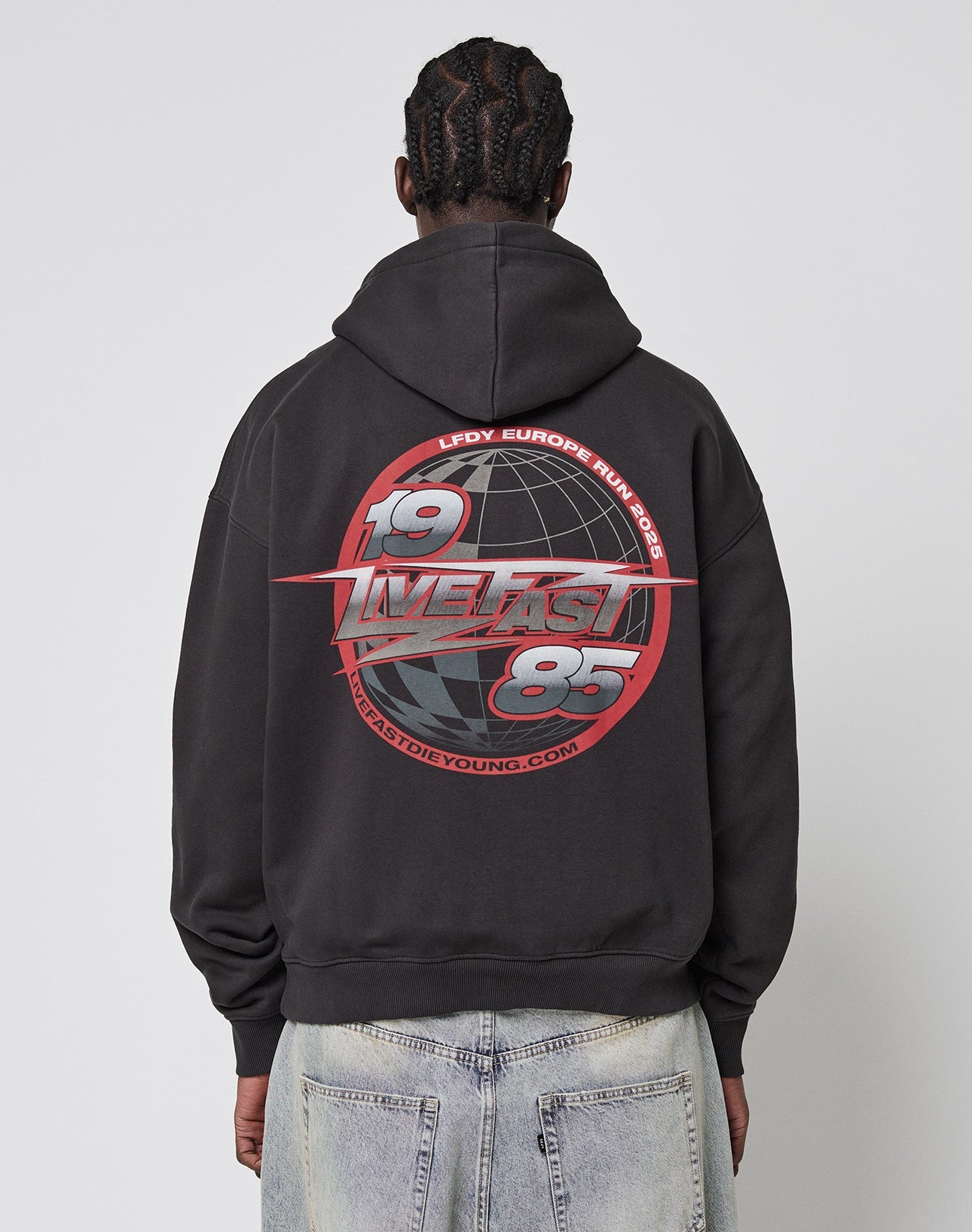 A person with braided hair wears the LFDY Globe Hooded by LIVE FAST DIE YOUNG, an oversized black hoodie featuring a bold red and white 1985 and Europe Run graphic with globe imagery, made from Baumwolle Polyester. Front and back print details included.