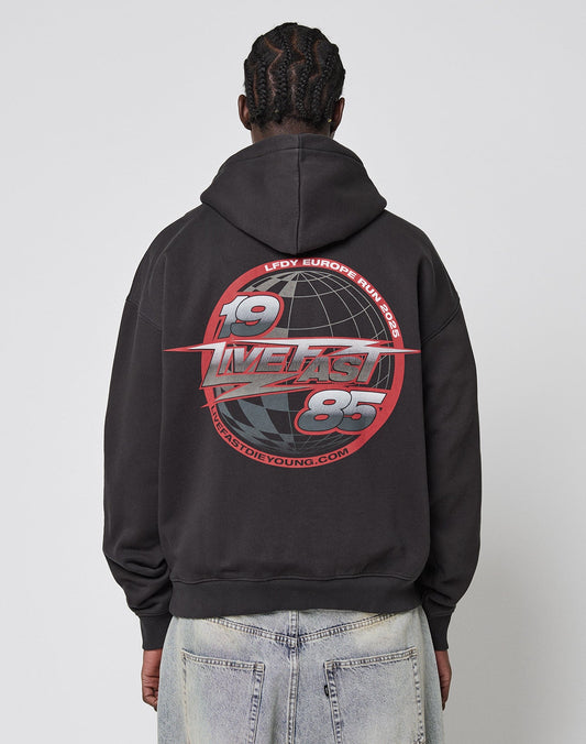 A person with braided hair wears the LFDY Globe Hooded by LIVE FAST DIE YOUNG, an oversized black hoodie featuring a bold red and white 1985 and Europe Run graphic with globe imagery, made from Baumwolle Polyester. Front and back print details included.