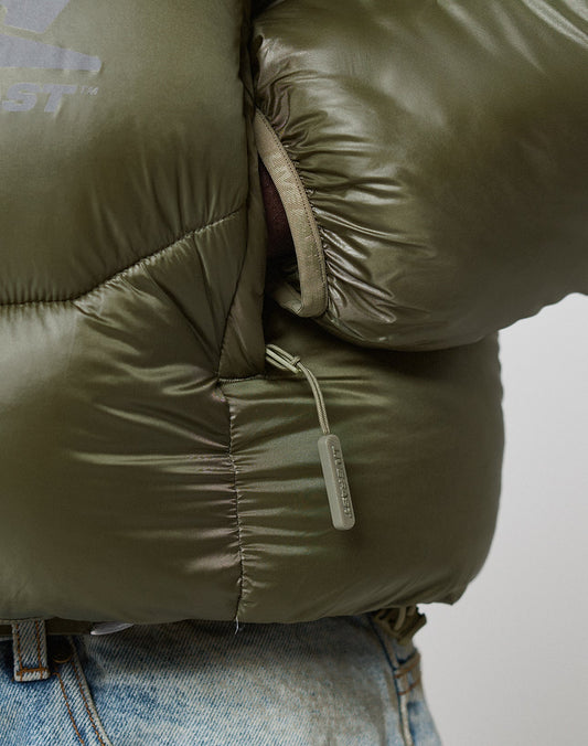 Close-up of a person in the Live Fast Puffer Jacket by LIVE FAST DIE YOUNG, featuring olive-green Nylon Obermaterial, reflektierender Print, a partially opened zippered underarm pocket showing dark fabric inside, and light blue jeans.