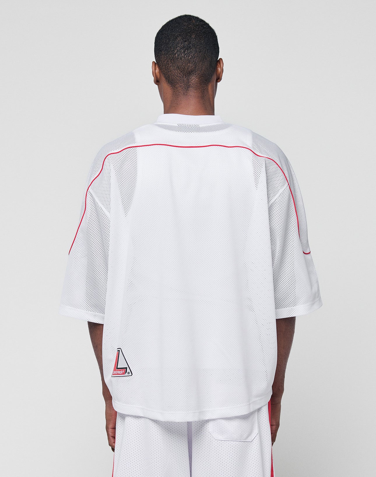 A person faces away from the camera, wearing the LIVE FAST DIE YOUNG Basic Mesh Jersey in white with red piping, a small geometric patch near the hem, V-neckline, and paired with matching white shorts.