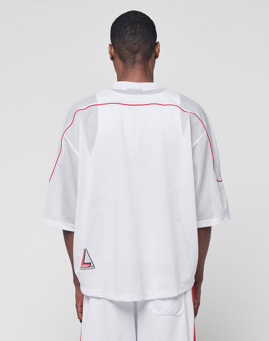 A person faces away from the camera, wearing the LIVE FAST DIE YOUNG Basic Mesh Jersey in white with red piping, a small geometric patch near the hem, V-neckline, and paired with matching white shorts.