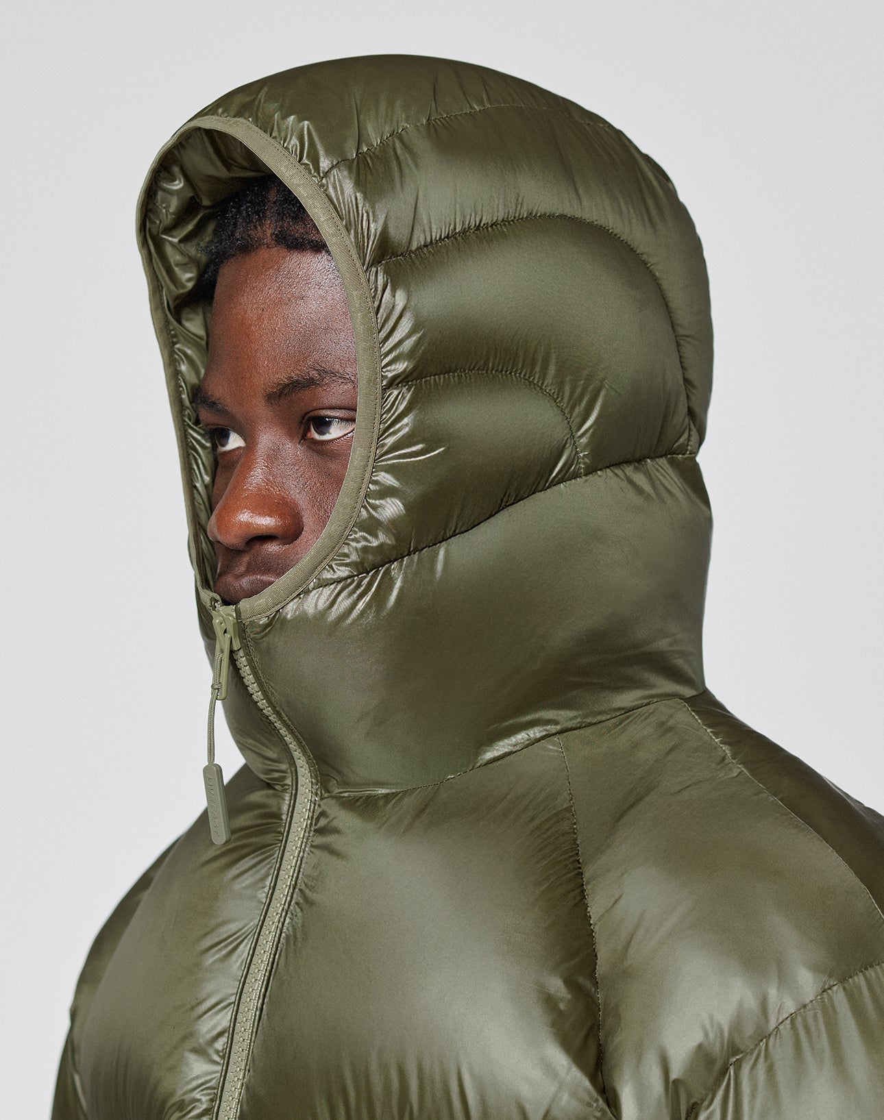 A person wearing the LIVE FAST DIE YOUNG Live Fast Puffer Jacket in olive green with oversized fit and nylon outer, hood up and zipped to the chin, gazes left against a plain light background.
