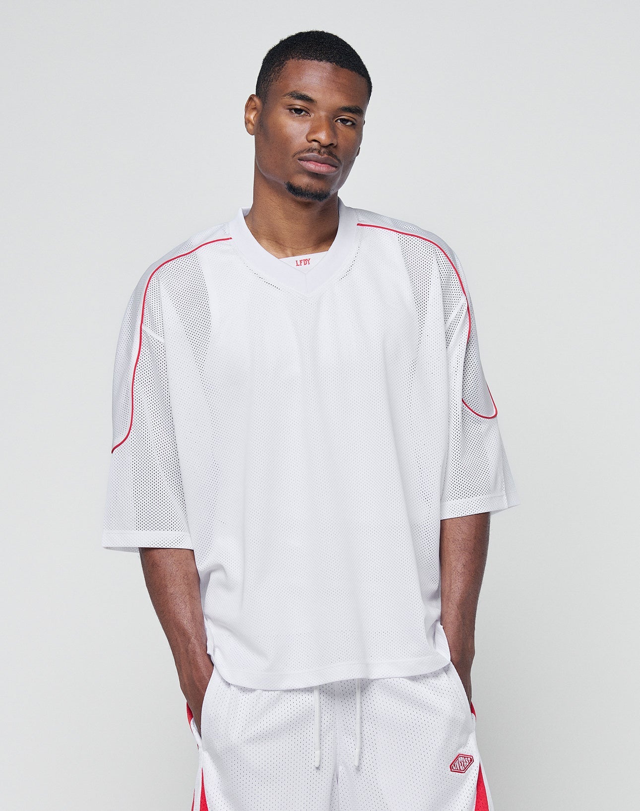 A man wears the LIVE FAST DIE YOUNG Basic Mesh Jersey— a loose-fitting, 100% polyester white shirt with red accents and matching shorts—standing against a plain light gray background, looking neutrally at the camera.