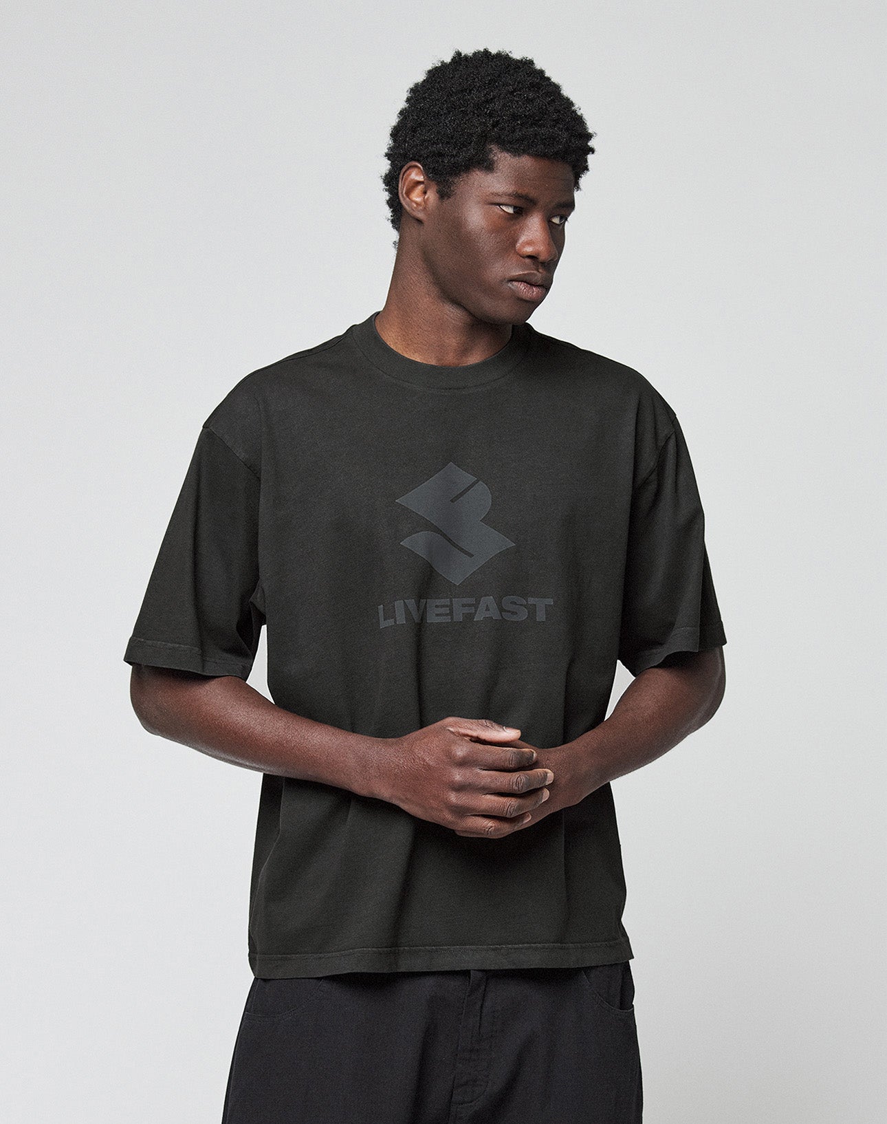 A person wears the Live Fast LF Tee by LIVE FAST DIE YOUNG, a black oversized t-shirt with a geometric logo and "LIVEFAST" front print. They stand against a light background, looking to the side with hands clasped.