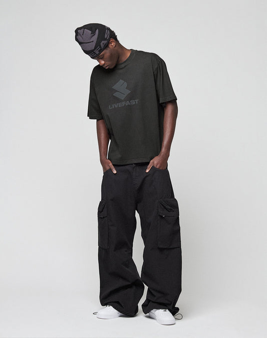 Someone stands against a plain light background wearing the black LIVE FAST DIE YOUNG Live Fast LF Tee (oversize fit, front print), loose black cargo pants, white sneakers, and a patterned black headwrap while looking down.