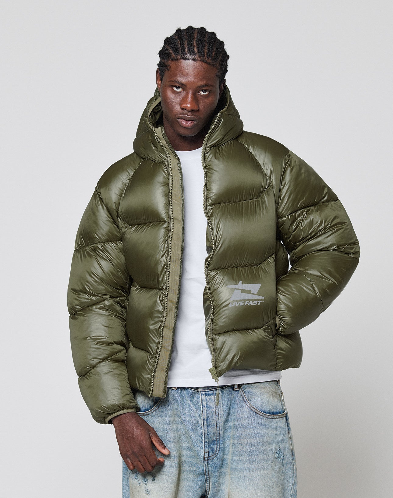 A man with braided hair wears a LIVE FAST DIE YOUNG Live Fast Puffer Jacket in olive green over a white T-shirt and faded blue jeans, standing against a plain light gray background.