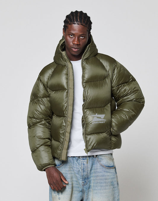 A man with braided hair wears a LIVE FAST DIE YOUNG Live Fast Puffer Jacket in olive green over a white T-shirt and faded blue jeans, standing against a plain light gray background.