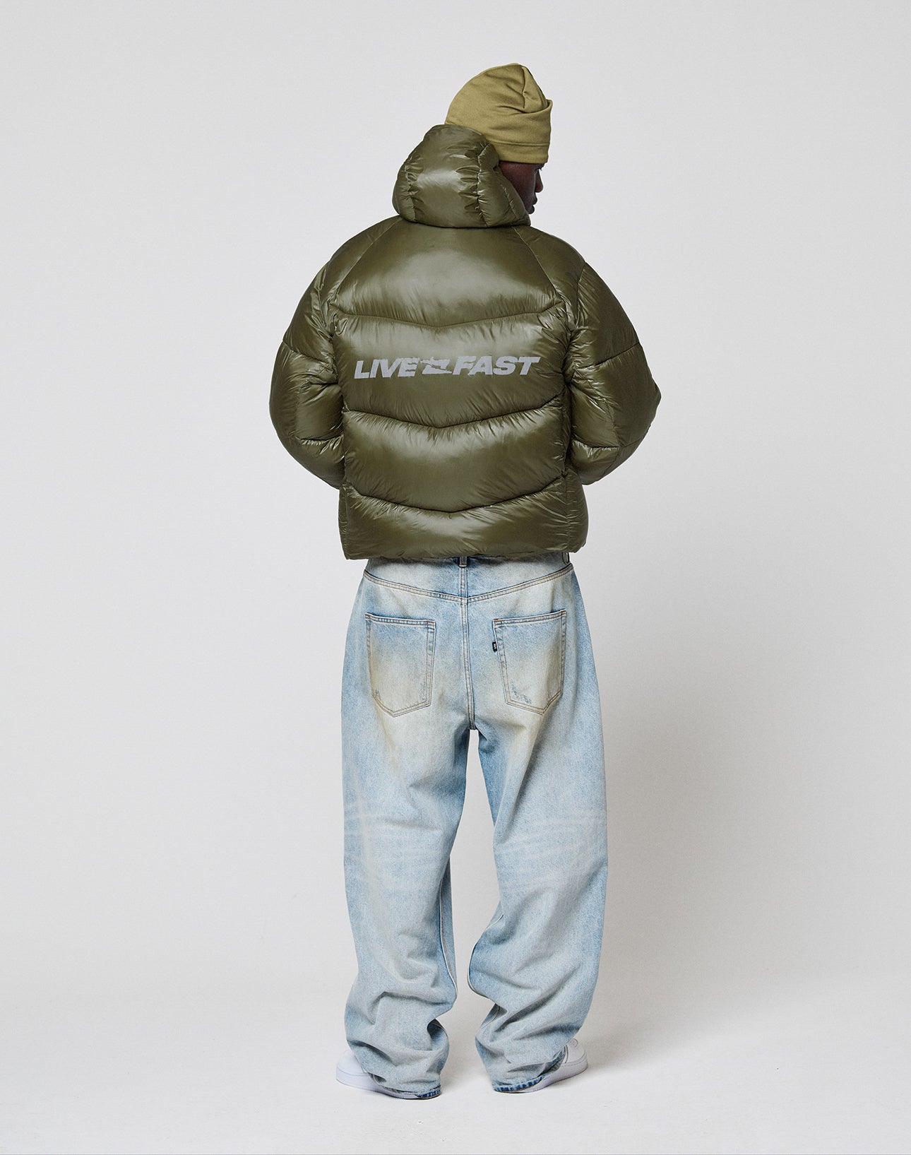 A person stands facing away in a LIVE FAST DIE YOUNG Live Fast Puffer Jacket with shiny olive nylon and a reflective LIVE FAST print on the back, paired with light blue loose jeans, a tan knit cap, and white sneakers against a plain background.