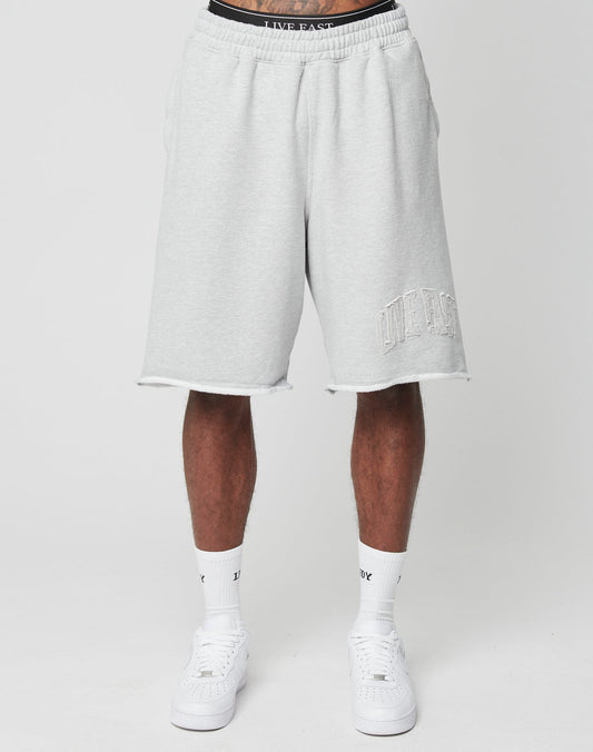 A person stands against a white background wearing LIVE FAST DIE YOUNG Ripped College Shorts in light gray with LIVE FAST embroidered on the left leg, styled with white socks and sneakers.