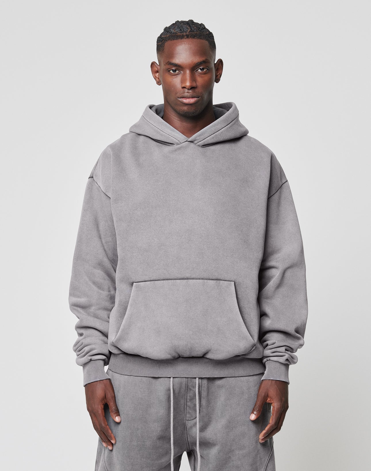 A man faces forward wearing a LIVE FAST DIE YOUNG Basic Blank Hooded, an oversized light gray cotton hoodie with a front pocket, paired with matching gray sweatpants. The background is plain and light-colored.