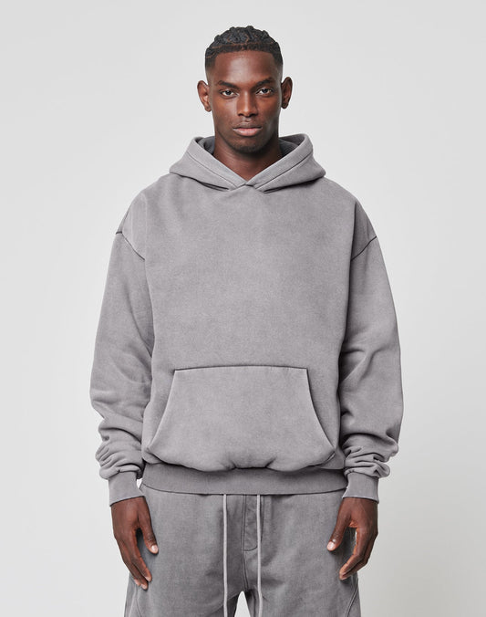 A man faces forward wearing a LIVE FAST DIE YOUNG Basic Blank Hooded, an oversized light gray cotton hoodie with a front pocket, paired with matching gray sweatpants. The background is plain and light-colored.