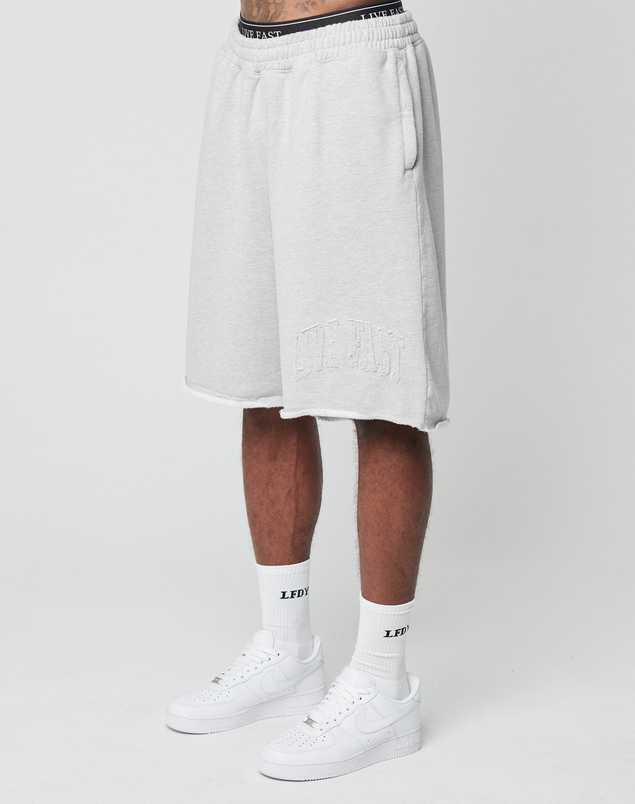 A person wears LIVE FAST DIE YOUNG Ripped College Shorts in light gray with an embroidered logo, knee length, paired with oversize white crew socks, white sneakers, and a black text waistband. The photo is on a plain white background.