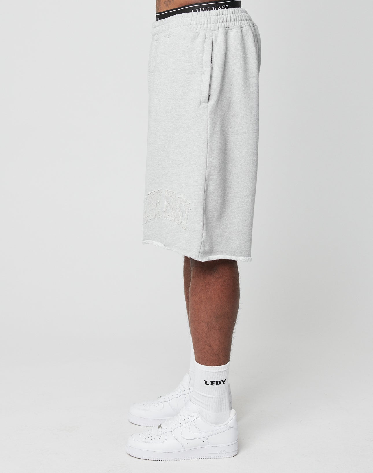 A person stands sideways in LIVE FAST DIE YOUNG Ripped College Shorts (light gray, knee-length, weitem Bein), white crew socks with “LEDY” text, and white Nike sneakers against a plain white background.