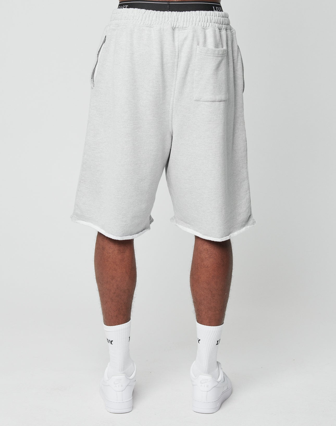 A person stands facing away, wearing LIVE FAST DIE YOUNG Ripped College Shorts in light gray with a back pocket, oversize fit and wide leg, paired with white crew socks and sneakers against a plain white background.