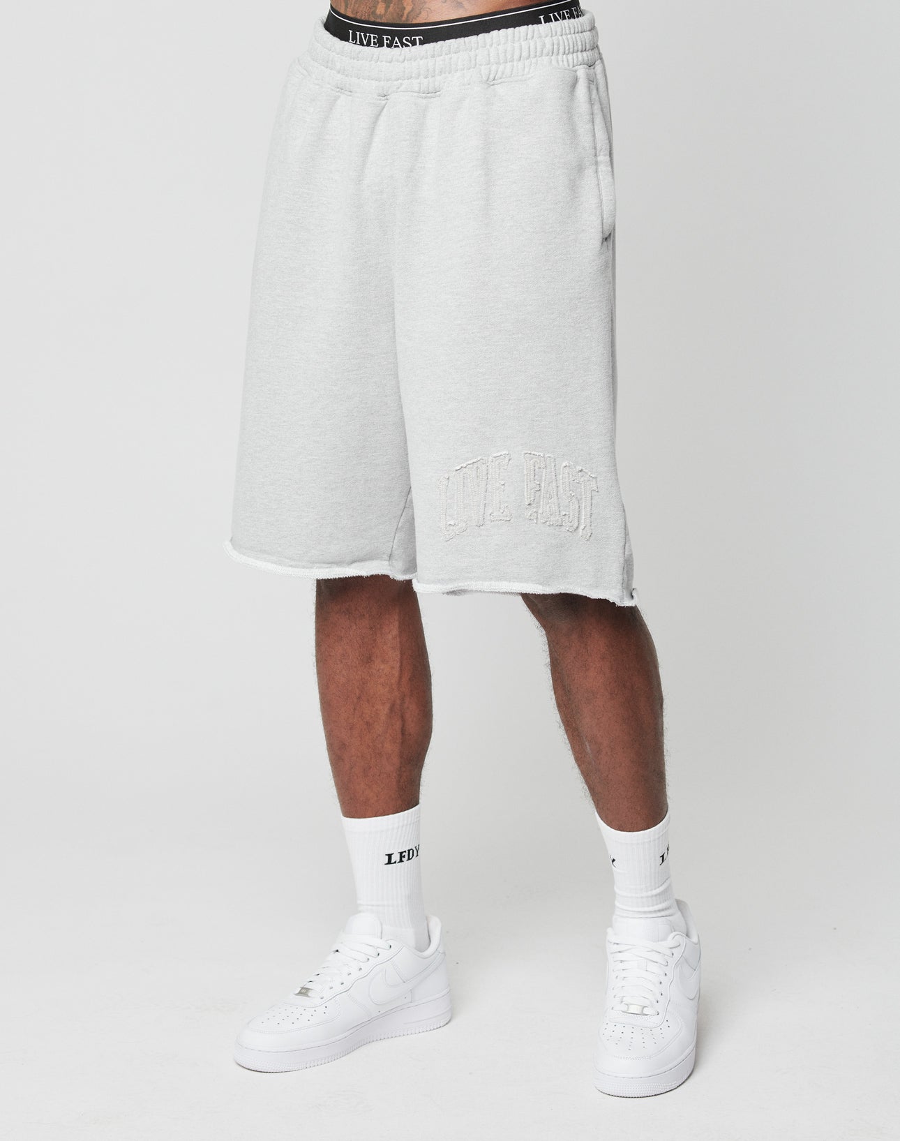A person in light gray, oversize fit Ripped College Shorts by LIVE FAST DIE YOUNG, white crew socks, and white sneakers stands against a plain white background.