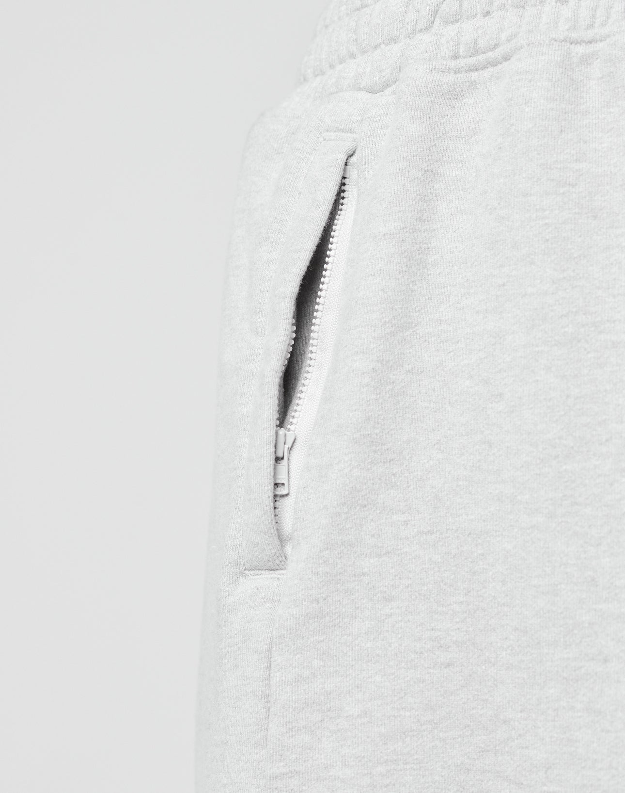 Close-up of a Ripped College Shorts pocket by LIVE FAST DIE YOUNG, featuring light gray fabric, a white zipper, and part of the elastic waistband. The shorts have an oversize fit and are set against a plain, light background.