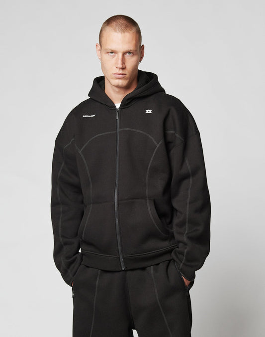 A person with short hair wears the LFDY Active Fleece Hooded Zip by LIVE FAST DIE YOUNG and matching black pants, standing with hands in pockets against a plain light gray background.