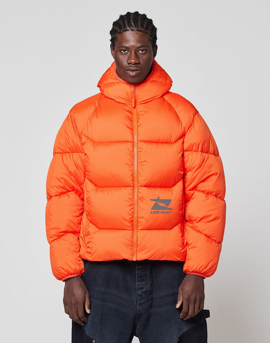 A man faces forward in the LIVE FAST DIE YOUNG Live Fast Puffer Jacket, an orange oversized style with a high collar and reflective "LIVE FAST" print plus graphic, paired with dark pants against a plain light background.| model