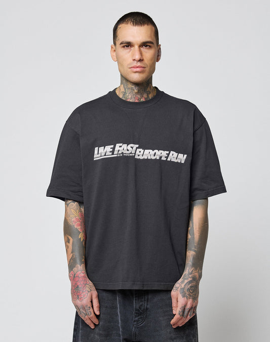 A tattooed man with short hair and a neutral expression wears the LFDY Europe Run Tee by LIVE FAST DIE YOUNG—an oversize black cotton t-shirt featuring LIVE FAST EUROPE RUN in white and gray. Waschhinweise included.