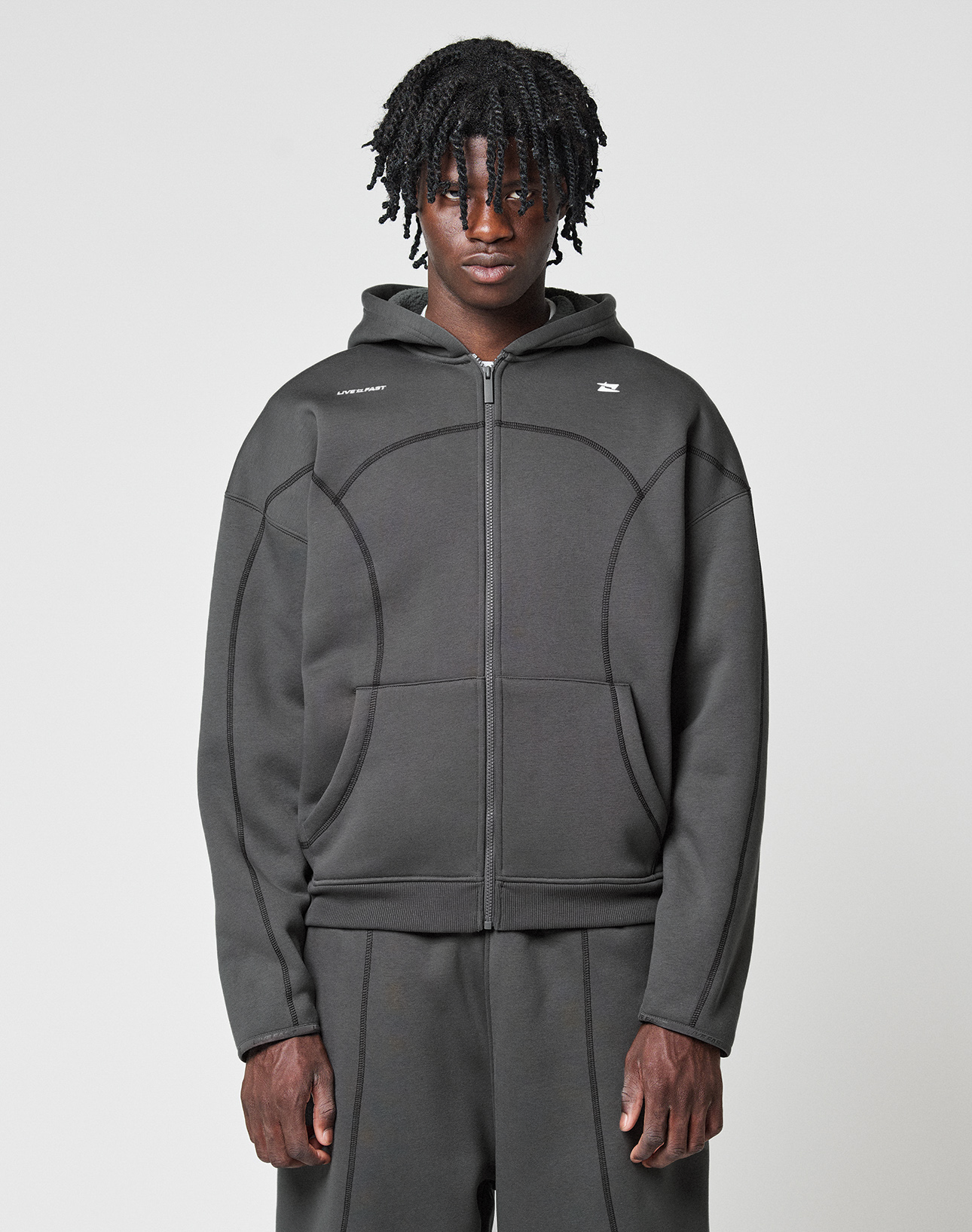 A person with medium-length curly hair wears the LFDY Active Fleece Hooded Zip by LIVE FAST DIE YOUNG in grey Oversize Fit, paired with matching pants, standing against a plain light background and looking directly at the camera with a neutral expression. | model