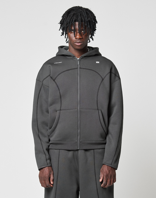 A person with medium-length curly hair wears the LFDY Active Fleece Hooded Zip by LIVE FAST DIE YOUNG in grey Oversize Fit, paired with matching pants, standing against a plain light background and looking directly at the camera with a neutral expression. | model