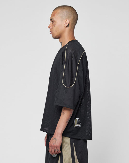 A person with a shaved head and visible tattoos stands in profile, wearing the LIVE FAST DIE YOUNG Basic Mesh Jersey in black with tan contrast piping and a geometric patch, paired with matching shorts, against a plain light gray background.