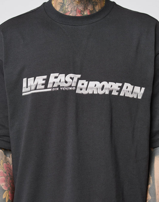 Someone wears the LFDY Europe Run Tee by LIVE FAST DIE YOUNG, an oversize black cotton t-shirt with bold white LIVE FAST DIE YOUNG EUROPE RUN lettering. Colorful tattoos are visible on their neck and arms. Follow care instructions for cotton.
