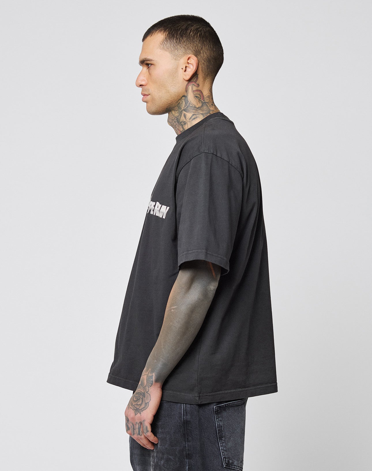 A tattooed man stands in profile, wearing the LIVE FAST DIE YOUNG LFDY Europe Run Tee and dark jeans against a plain light grey background. Care instructions are on the label.