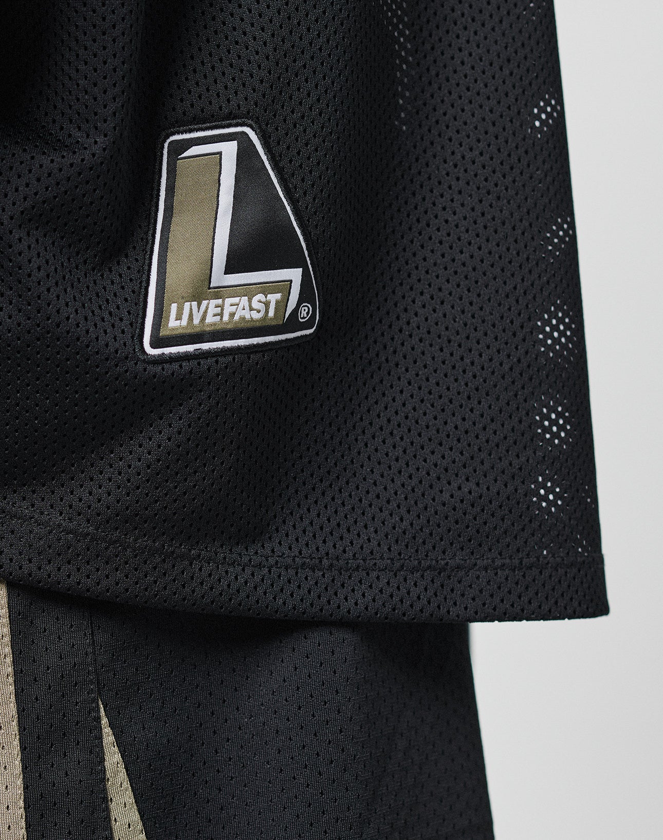 Close-up of the black Basic Mesh Jersey by LIVE FAST DIE YOUNG, featuring Kontrast Piping and a patch with a bold white L and LIVEFAST text on a gold and black background.