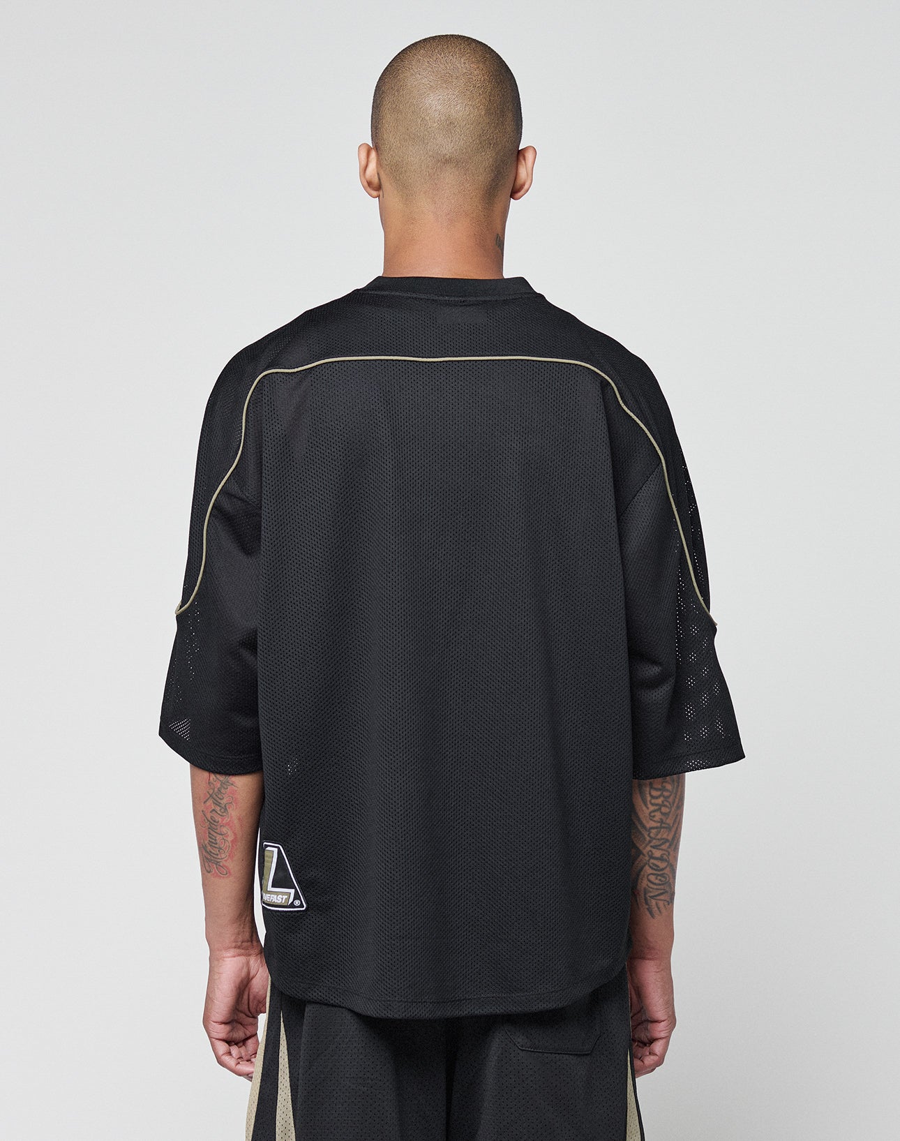 A person with a shaved head faces away, wearing the LIVE FAST DIE YOUNG Basic Mesh Jersey in black with Kontrast Piping and a small white logo near the hem. The oversize fit accentuates their tattooed arms against a plain, light background.