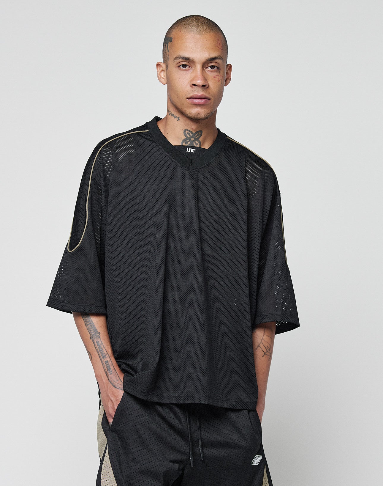 A person with short hair and visible tattoos, wearing the LIVE FAST DIE YOUNG Basic Mesh Jersey in oversize black with Kontrast piping and matching black shorts with beige accents, stands against a plain light gray background.