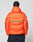 A person with braided hair wears the Live Fast Puffer Jacket by LIVE FAST DIE YOUNG—an oversize, bright orange jacket with bold blue LIVE FAST lettering and a reflektierender Print—standing against a plain, light background.