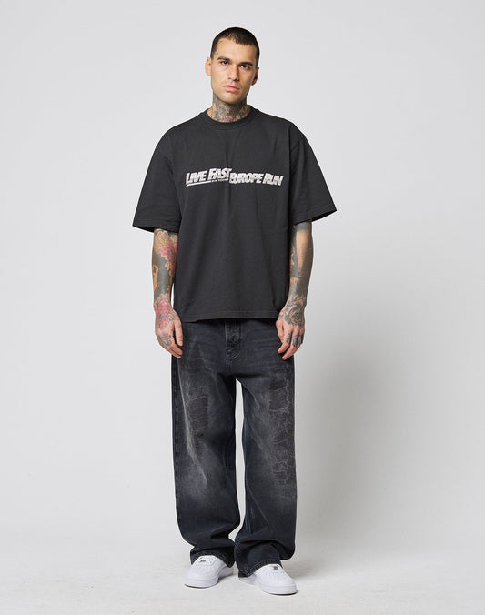 A tattooed man stands facing forward in a LIVE FAST DIE YOUNG LFDY Europe Run Tee, loose black T-shirt, baggy black jeans, and white sneakers against a plain light background.