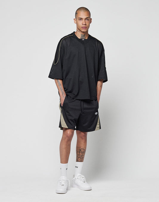 A man with tattoos stands against a plain white background wearing a black LIVE FAST DIE YOUNG Basic Mesh Jersey, black shorts with beige contrast piping, white crew socks, and white sneakers. He has his hands in his pockets and a neutral expression.