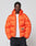 A person wears the LIVE FAST DIE YOUNG Live Fast Puffer Jacket in orange with an oversize fit, water-repellent finish, small graphic, and "LIVE FAST" text on the left, styled over a white t-shirt and black pants against a light background.