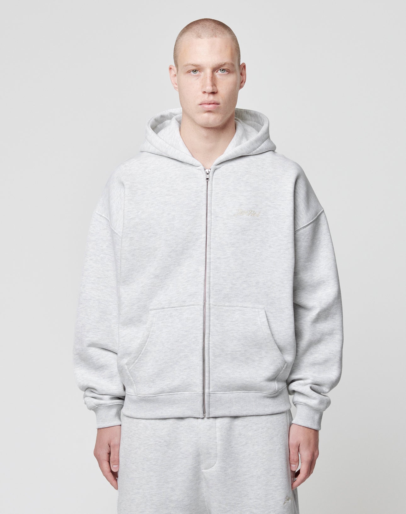 A person with a shaved head wears the LIVE FAST DIE YOUNG Basic Script Hooded Zip and matching sweatpants in light gray, both with a subtle Logo-Stick, standing against a white background and facing the camera neutrally.