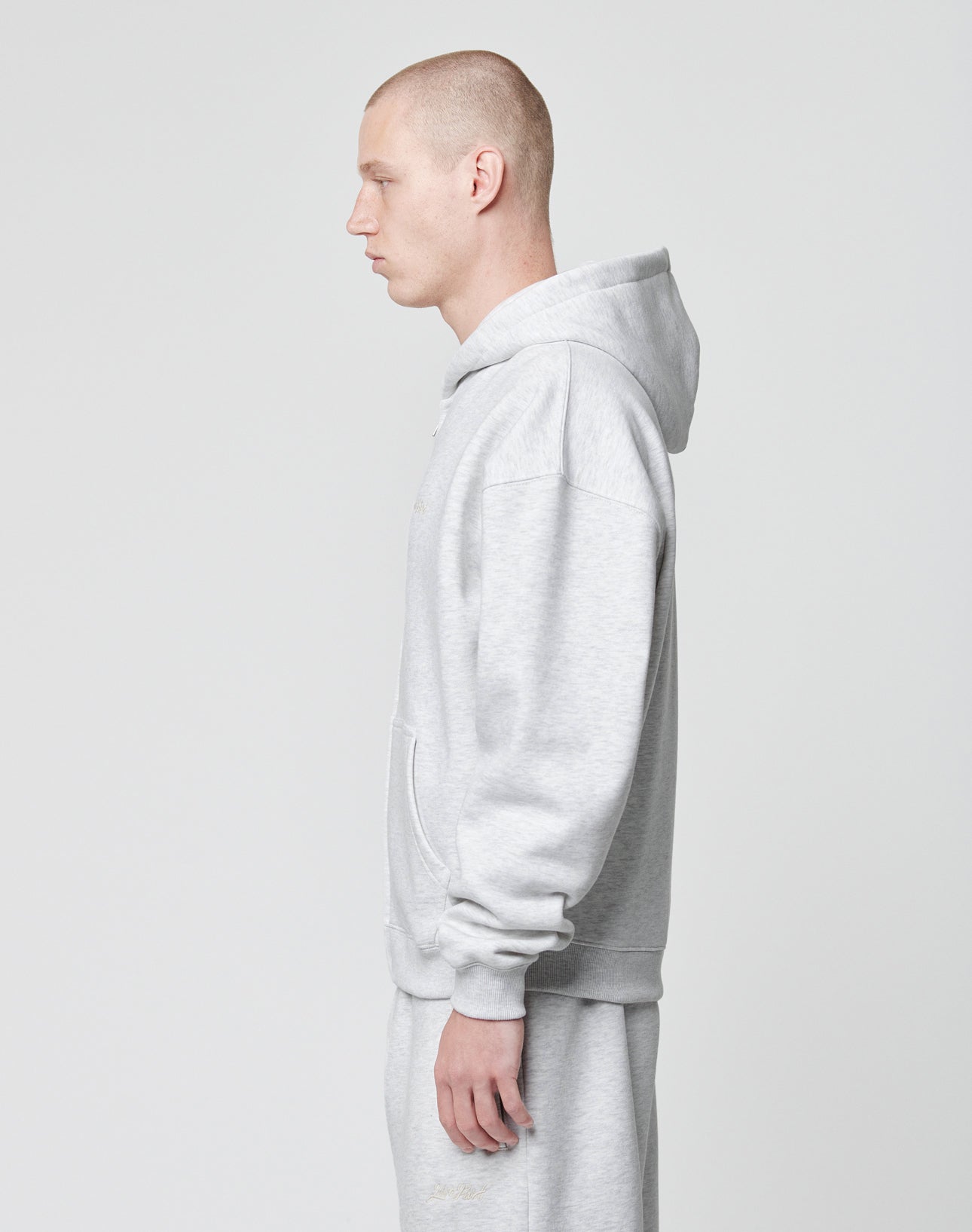 A person with a shaved head models the LIVE FAST DIE YOUNG Basic Script Hooded Zip and matching sweatpants in light gray Baumwolle-Polyester, standing in profile against a plain white background.