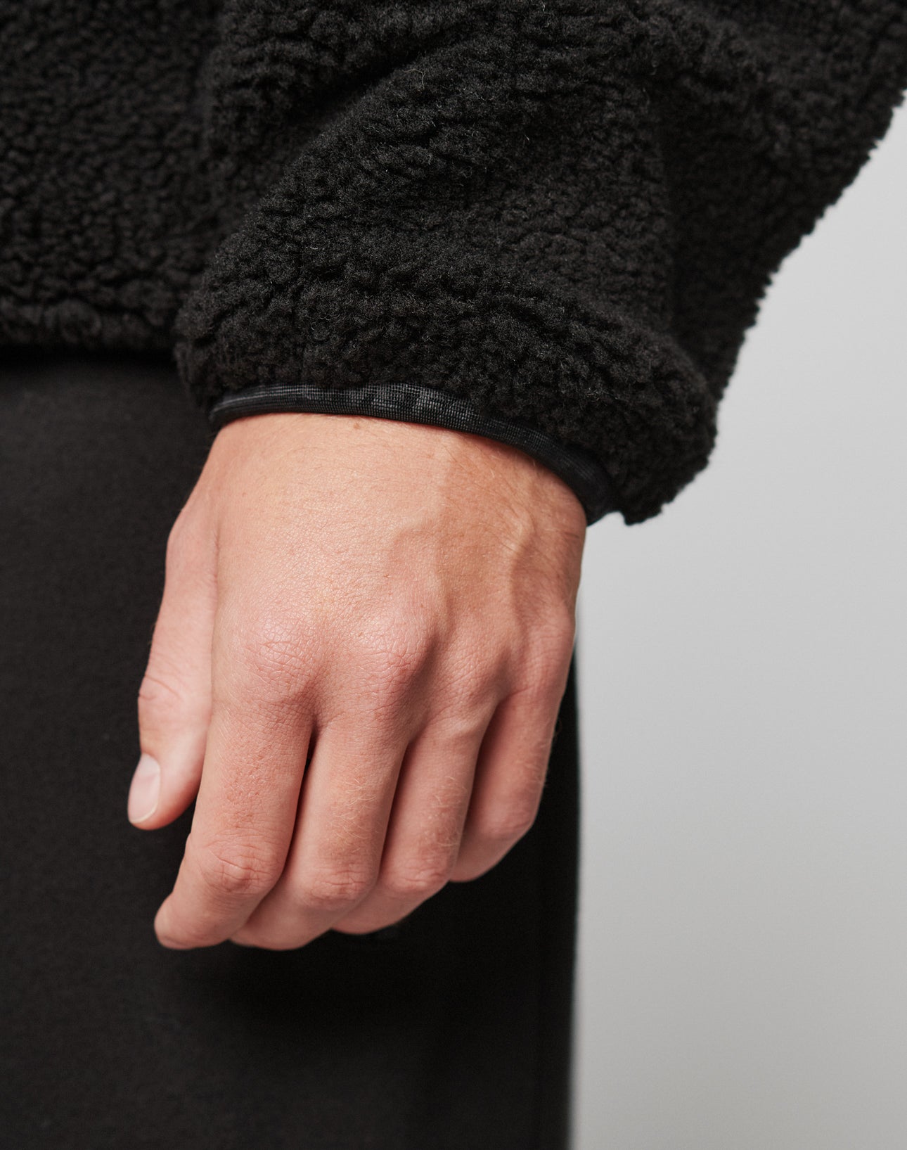 A close-up of a person’s left hand relaxed at their side, wearing the LIVE FAST DIE YOUNG Live Fast Teddy Jacket in black with textured fabric and subtle embossed branding, set against a plain, light-colored background.