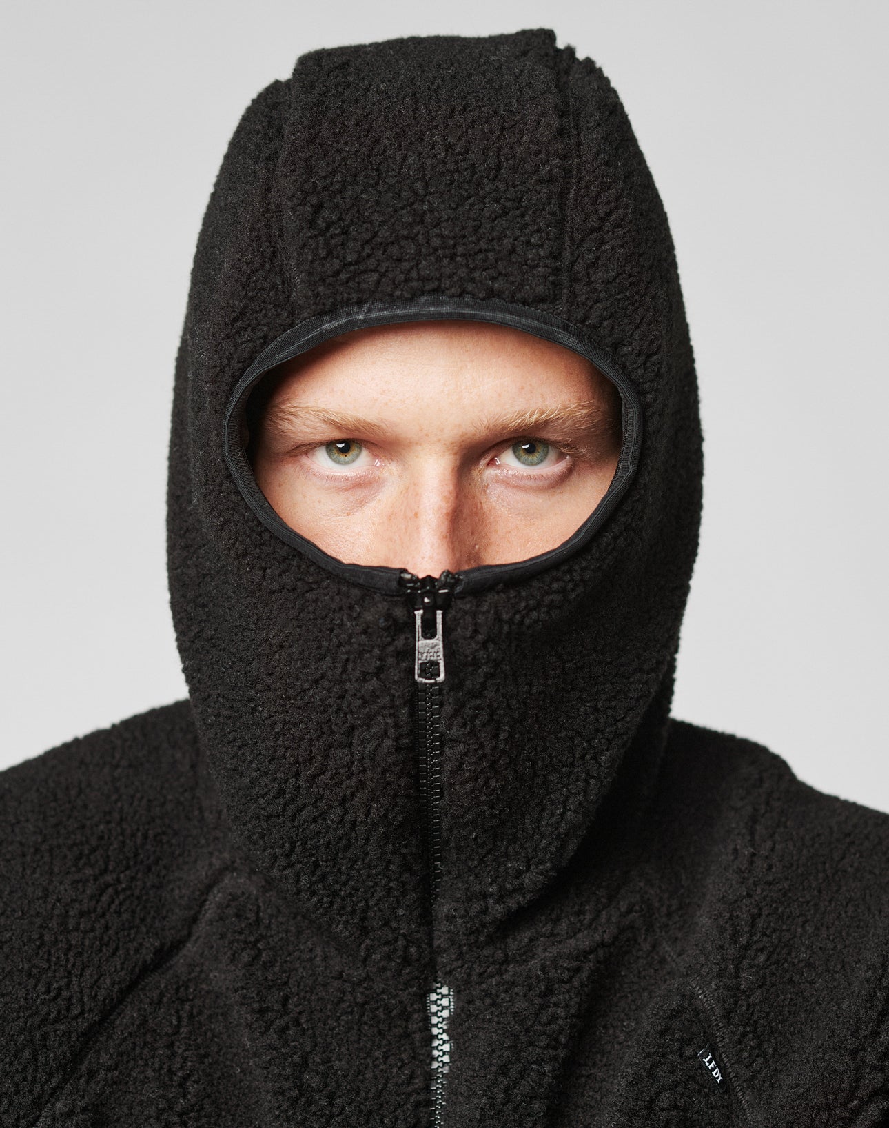 A person in a LIVE FAST DIE YOUNG Live Fast Teddy Jacket, with hood zipped to show just their eyes, nose, and forehead, stands against a plain light grey background. Subtle embossed branding adds a refined touch.