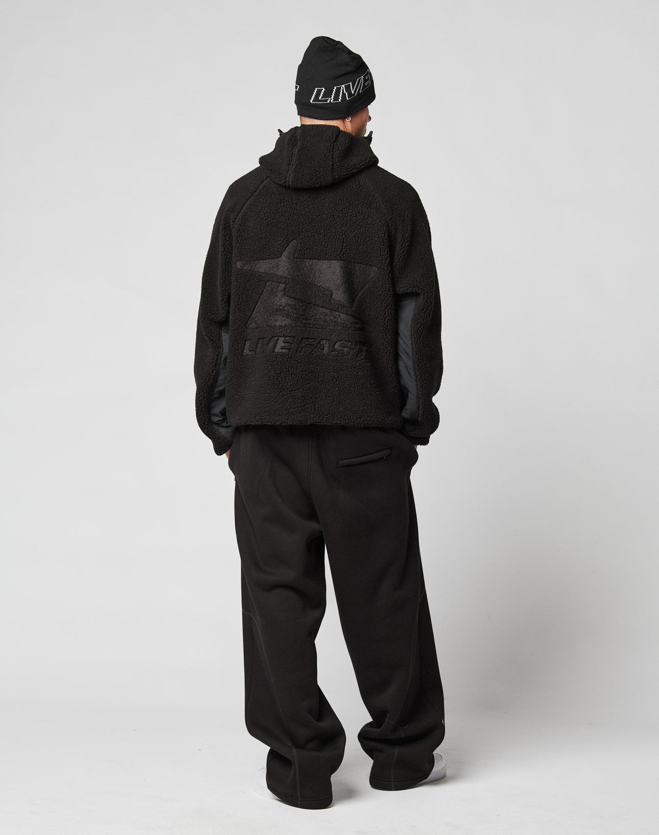A person faces away, wearing the LIVE FAST DIE YOUNG Live Fast Teddy Jacket with embossed branding and LIVE FAST graphic on the back, paired with a black beanie and loose black pants against a plain white background.