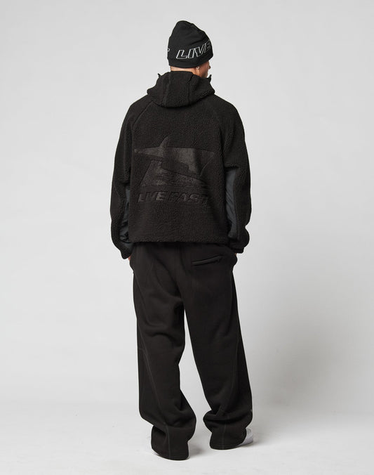 A person faces away, wearing the LIVE FAST DIE YOUNG Live Fast Teddy Jacket with embossed branding and LIVE FAST graphic on the back, paired with a black beanie and loose black pants against a plain white background.