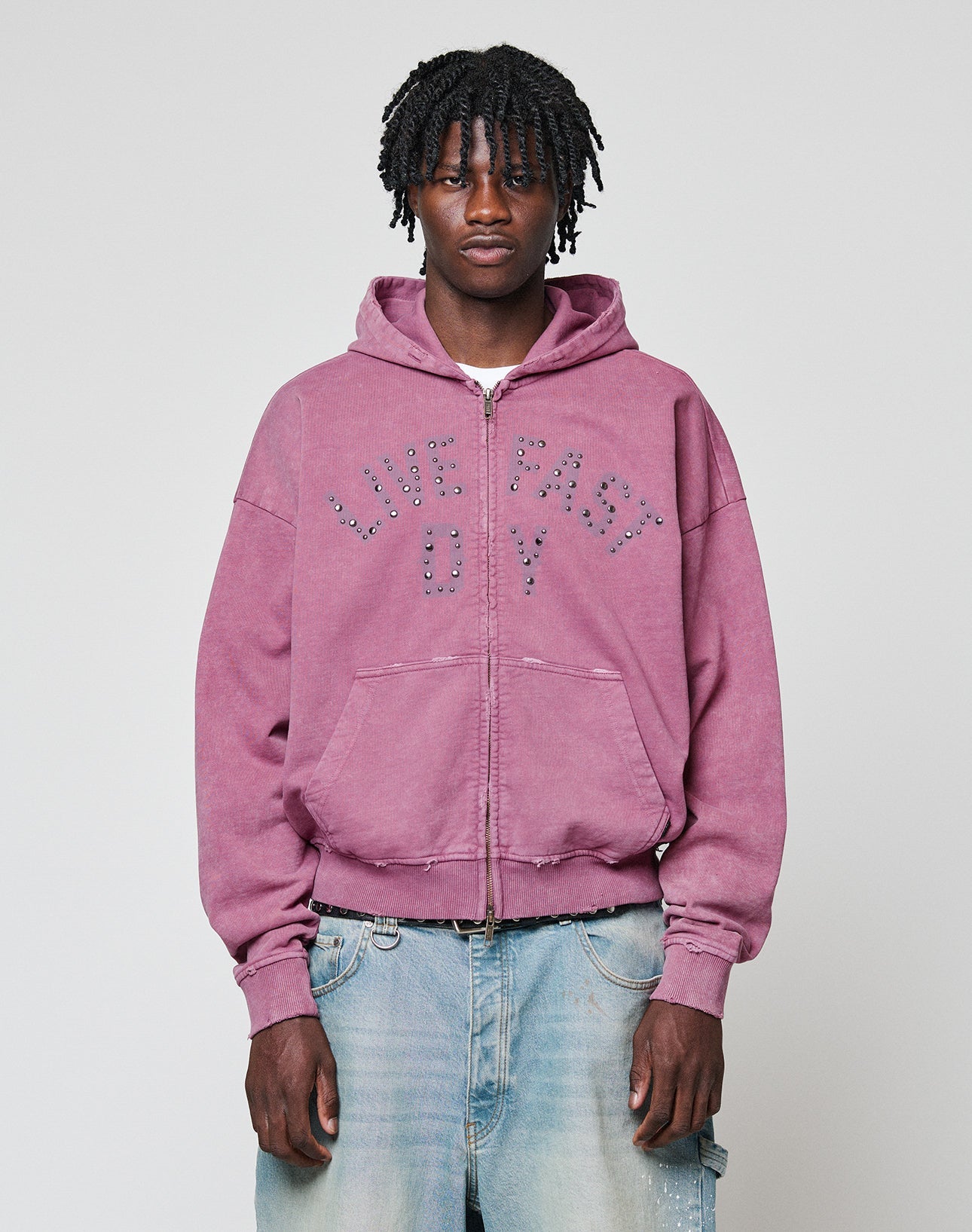 A young man with short dreadlocks wears the Live Fast Rivet Hooded Zip by LIVE FAST DIE YOUNG—a light purple hoodie featuring studded lettering and a handmade spray effect—paired with faded blue jeans, against a plain light gray background.