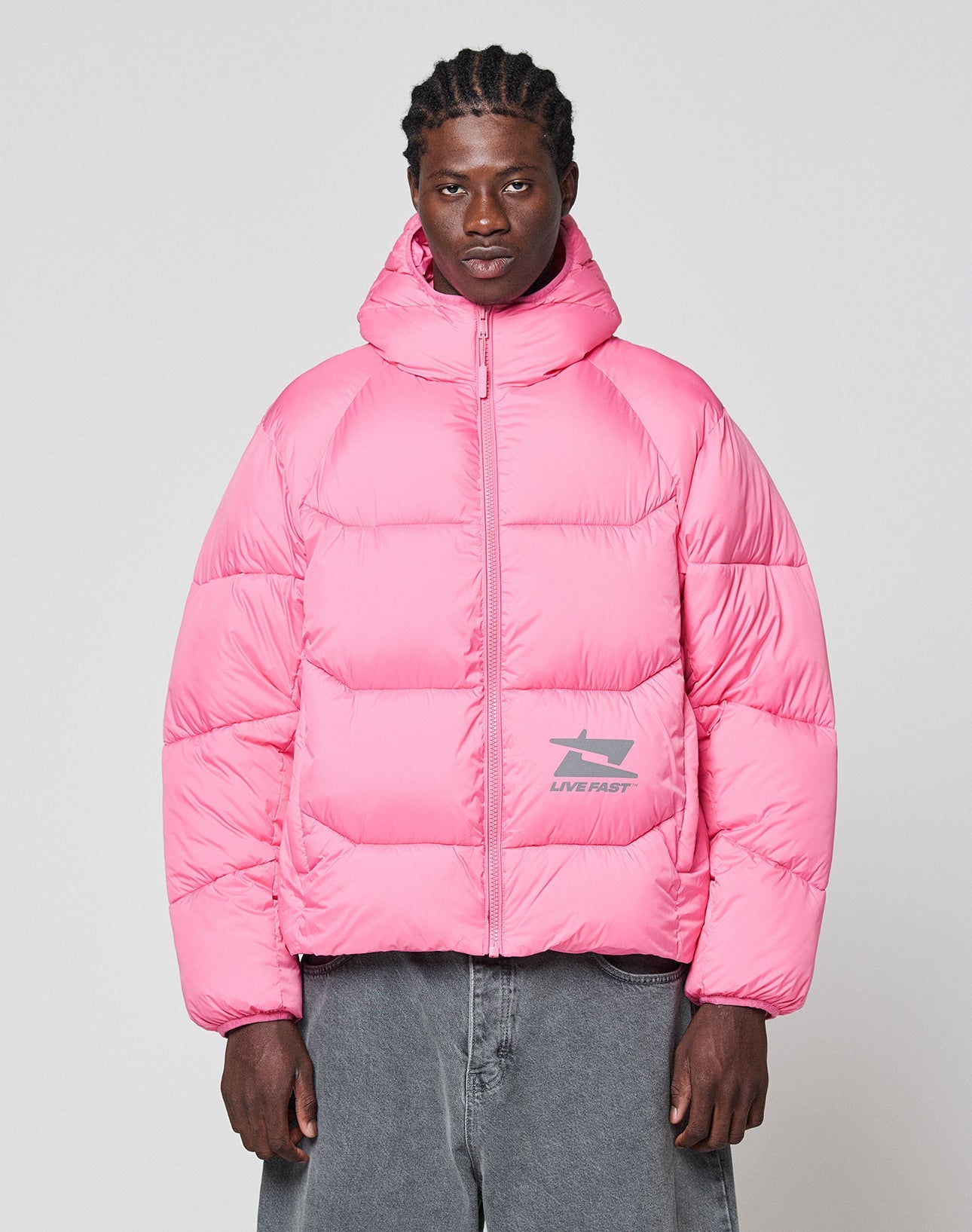 Wearing the LIVE FAST DIE YOUNG Live Fast Puffer Jacket in oversize bright pink nylon with hood and reflective shark logo print, a person with braided hair and loose gray pants stands against a light gray background.
