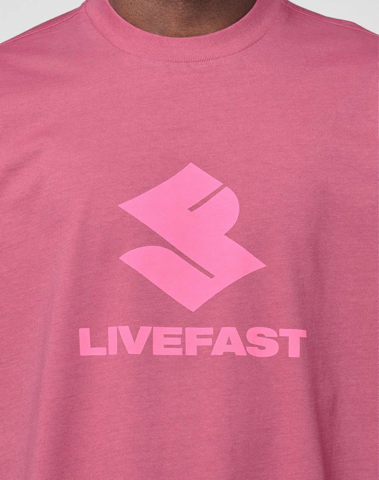 A close-up of a person wearing the Live Fast LF Tee by LIVE FAST DIE YOUNG, featuring a bold abstract front logo above "LIVEFAST" in pink block letters. The oversize fit adds comfort.