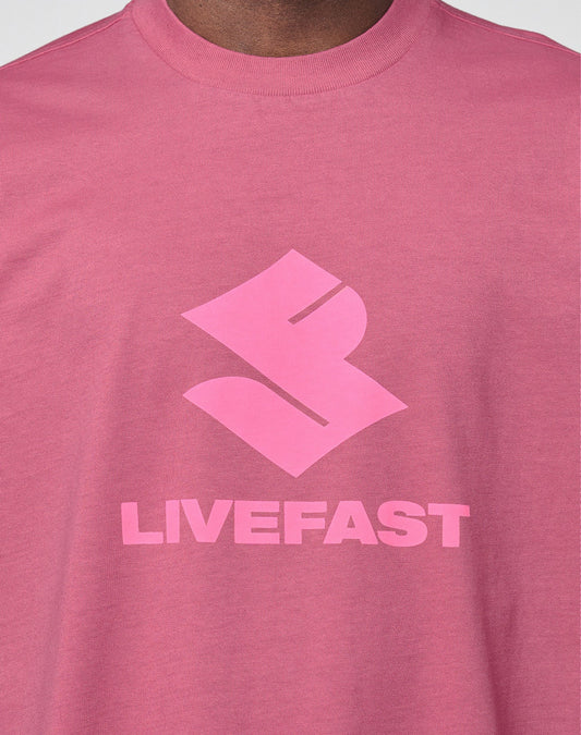 A close-up of a person wearing the Live Fast LF Tee by LIVE FAST DIE YOUNG, featuring a bold abstract front logo above "LIVEFAST" in pink block letters. The oversize fit adds comfort.
