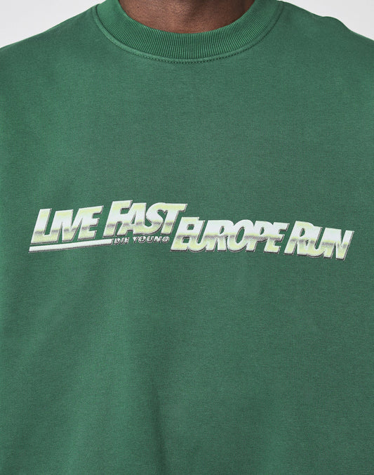 A person wears the LIVE FAST DIE YOUNG LFDY Europe Run Sweater, an oversize green sweatshirt with LIVE FAST EUROPE RUN printed in bold white and green gradient letters across the chest.