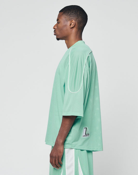 A man stands in profile wearing the LIVE FAST DIE YOUNG Basic Mesh Jersey and matching shorts in mint green with white contrast piping. The 100% polyester shirt features a small white and black patch on the lower back, set against a plain white background.