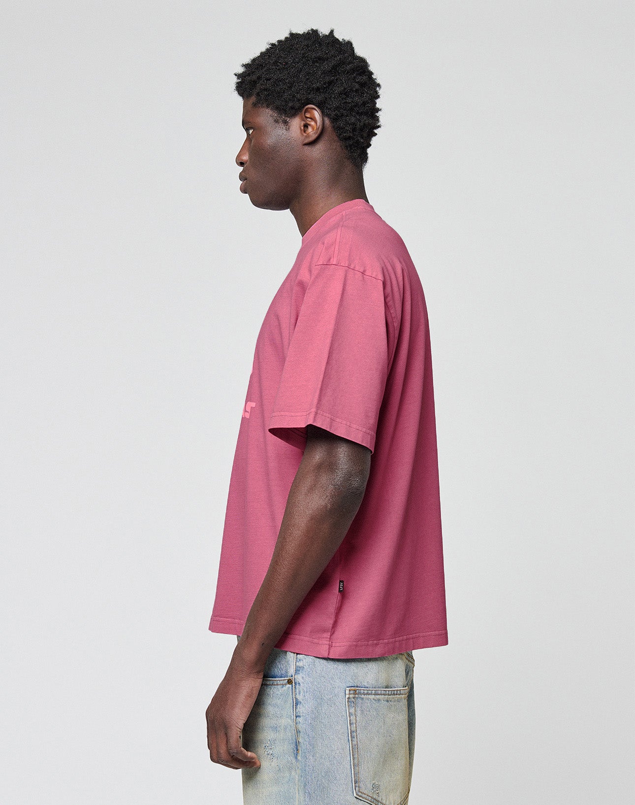 A man in profile faces left, wearing a LIVE FAST DIE YOUNG Live Fast LF Tee in mauve with an oversize fit and light blue jeans, standing against a plain light gray background.